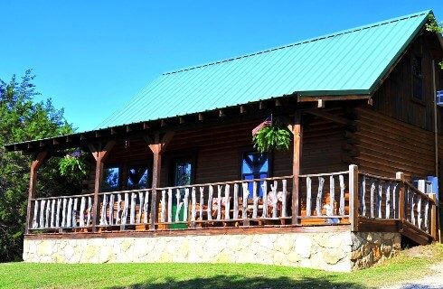 Wood log cabin with green roof, front porch with railing hanging fern baskets