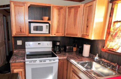 Corner of a kitchen with wood cabinets, stove, microwave, sink and bright window