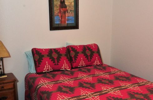 Small bedroom with queen bed with a red quilt and side table with a lamp