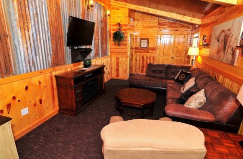 Cabin living room with tin and wood walls, brown leather sectional sofa, chair and TV hung over dresser