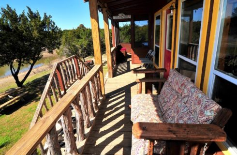 Front porch of a cabin with love seat and chairs overlooking a pond and trees