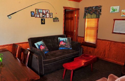 Living room of a cabin with brown couch, table and chairs, coffee table and doorway into a bedroom