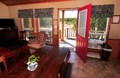 Interior of a cabin with dark wood kitchen table, velvet chair, dresser with TV, windows with valences and doorway open to a front porch