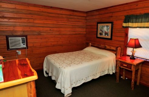 Cabin bedroom with queen bed, white quilt, side table with lamp, dresser and a shaded window