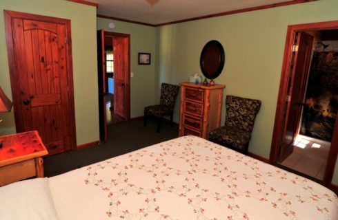 Bedroom with green walls, queen bed in white quilt, dresser, two chairs and doorways into bathroom and hallway