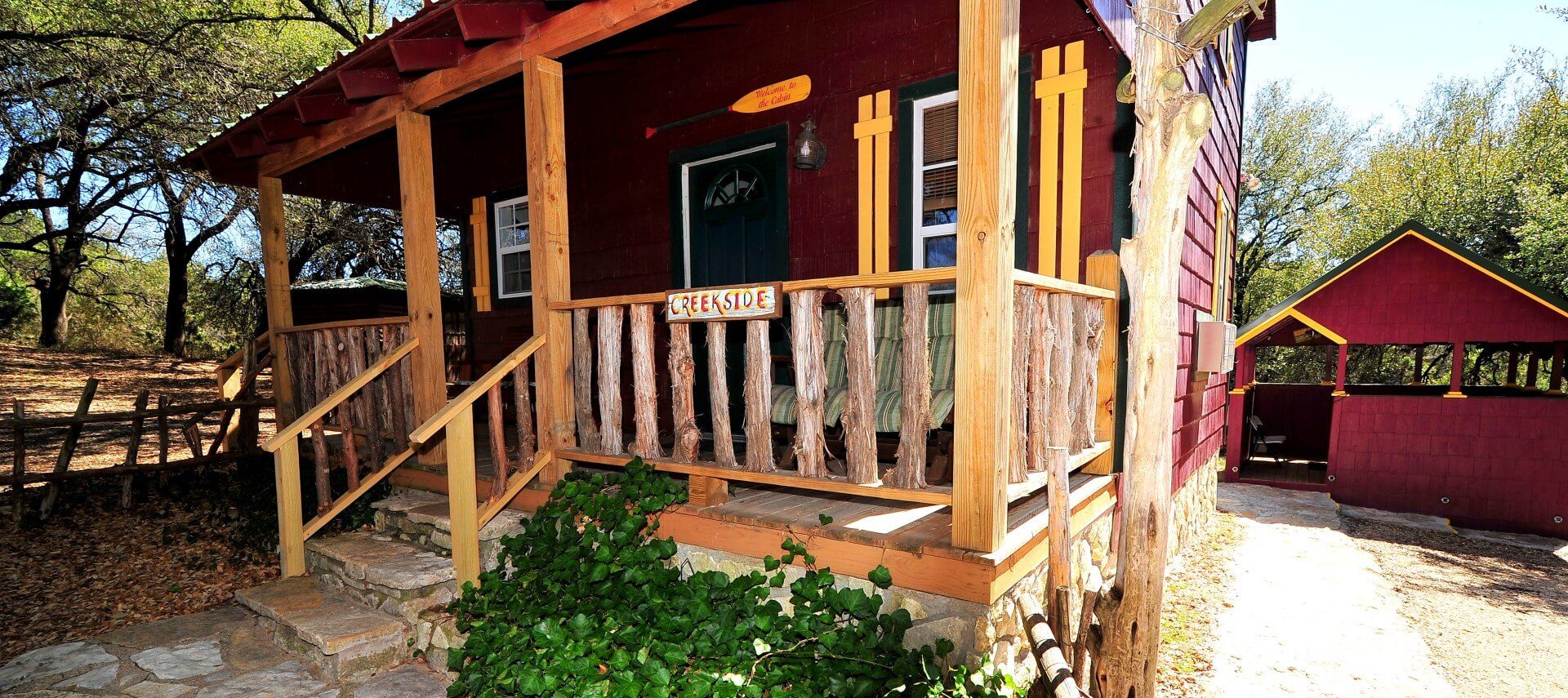 Small red cabin with yellow trim, front porch and red building behind at the end of a walkway