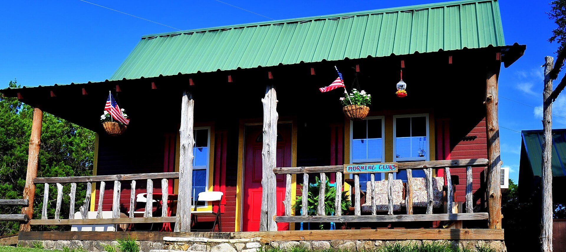 Quaint brown cabin with yellow trim, green roof, front porch and hanging flower baskets