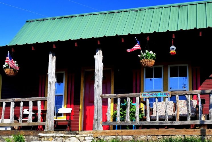 Quaint brown cabin with yellow trim, green roof, front porch and hanging flower baskets