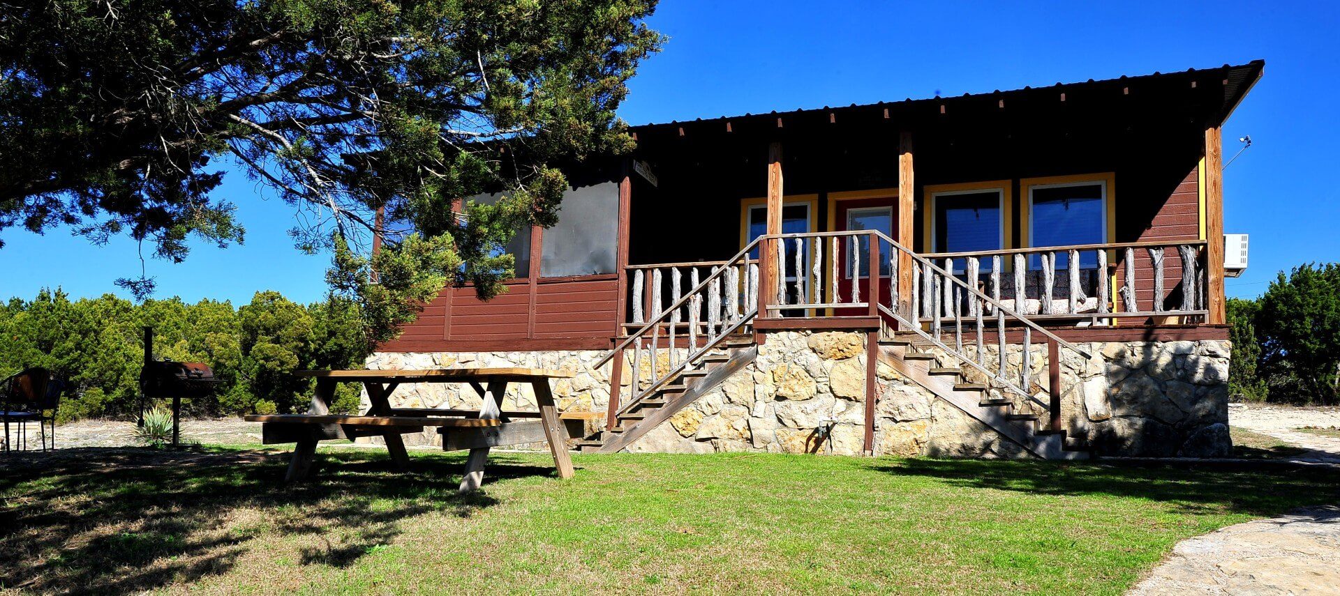 Brown cabin with yellow trim and stone detail, front porch and picnic table on lawn by a large tree