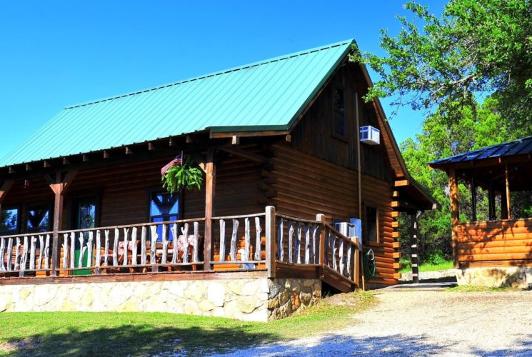 Wood log cabin with green roof, front porch with railing and side building with open air windows